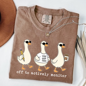 May include: Brown Comfort Colors t-shirt featuring three cartoon ducks wearing sunglasses. One duck holds a pencil, another a shirt with "YOU GOT THIS", and the last a coffee cup. The text "off to actively monitor" is printed below.