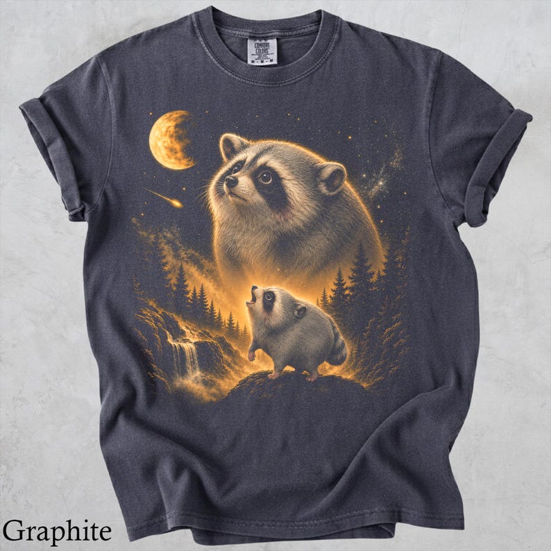 May include: A charcoal gray t-shirt featuring a graphic print of a large raccoon looking up at a glowing moon and a smaller raccoon standing on a rocky cliff near a waterfall, surrounded by a forest landscape with a shooting star in the night sky.