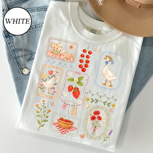 Pode incluir: Camiseta branca com um design l&uacute;dico com um ganso com chap&eacute;u azul, morangos, uma chaleira e flores. A camisa tem uma etiqueta Comfort Colors e est&aacute; colocada em cima de jeans azuis.
