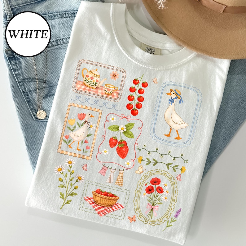 Pode incluir: Camiseta branca com um design l&uacute;dico com um ganso com chap&eacute;u azul, morangos, uma chaleira e flores. A camisa tem uma etiqueta Comfort Colors e est&aacute; colocada em cima de jeans azuis.