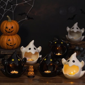 Puede incluir: Decoraciones de Halloween de cerámica. La imagen presenta calabazas naranjas apiladas con caras talladas y candelabros con forma de fantasma en blanco y negro. Una pequeña pila de figuras de calabaza y velas encendidas completan el ambiente espeluznante.