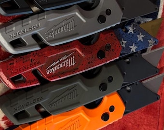 May include: Collection of Milwaukee Fastback utility knives in various colors, including blue, black, red, green, pink, gray, and orange. Each knife features a retractable blade and the "FASTBACK" and "Milwaukee" logos.