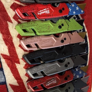 May include: Collection of Milwaukee FASTBACK utility knives in various colors, including blue, black, red, green, pink, gray, and orange. Each knife features a blade and the Milwaukee logo.