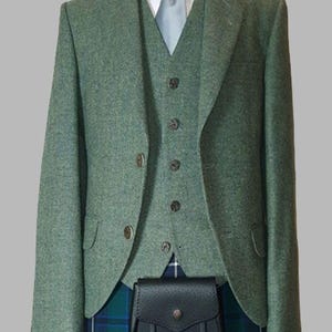 Op de afbeelding: Een groene tweed jas, vest en kilt outfit. De jas en het vest hebben donkerbruine knopen. Een wit overhemd en een lichtblauwe stropdas zijn zichtbaar. Een zwarte leren sporran met kwastjes hangt aan de kilt. De kilt heeft een blauw, groen en wit ruitpatroon.
