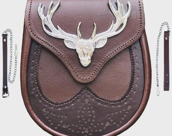 Può includere: Un sporran in pelle marrone con un'emblema a testa di cervo argentata. Lo sporran ha una chiusura a patta e cuciture decorative. Due cinturini in pelle con dettagli a catena pendono dai lati.