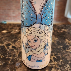 May include: A white tumbler with a cartoon illustration of Elsa from Frozen, a large blue bow, and castle details. The design includes the names of Disney characters like Tinker Bell and Pinocchio. The tumbler has a silver lid.