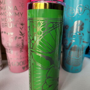 May include: A collection of insulated tumblers in various colors. The central tumbler is green with a gold floral design. Other tumblers are pink and teal, with white lids and various text and graphic designs.