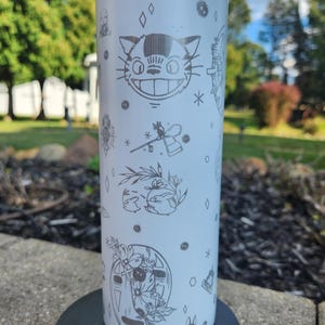 May include: White tumbler with a blue lid, featuring etched illustrations of characters and symbols from a popular animated film. The design includes a smiling cat face, stars, and other whimsical elements. The tumbler is cylindrical and stands on a black base.