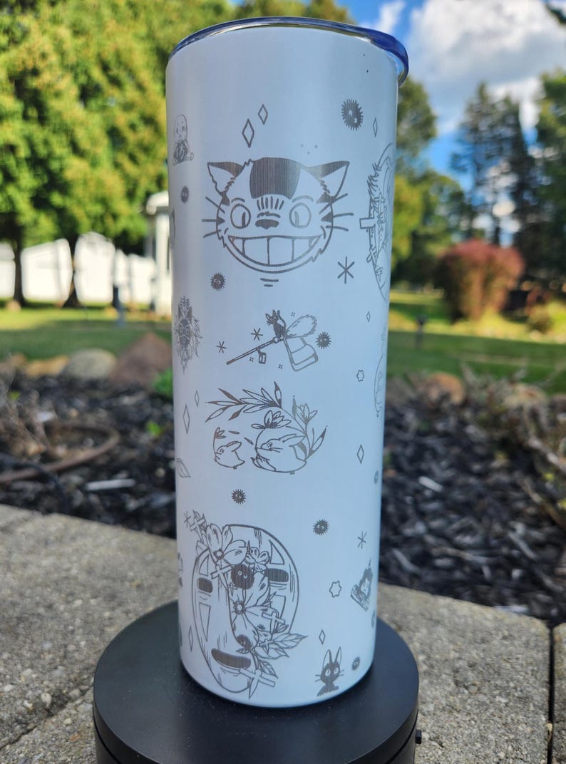 May include: White tumbler with a blue lid, featuring etched illustrations of characters and symbols from a popular animated film. The design includes a smiling cat face, stars, and other whimsical elements. The tumbler is cylindrical and stands on a black base.