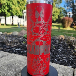 Puede incluir: Un vaso rojo de acero inoxidable con un diseño de Deadpool grabado con láser. El diseño incluye la imagen del personaje, espadas cruzadas y la palabra "DEADPOOL". El vaso tiene una tapa plateada y está diseñado para mantener las bebidas frías o calientes.