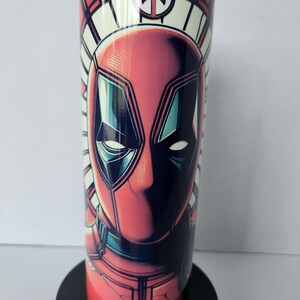 Puede incluir: Un vaso rojo y negro con una ilustraci&oacute;n detallada de la cara de Deadpool. El dise&ntilde;o incluye un patr&oacute;n geom&eacute;trico blanco y rojo alrededor de la parte superior y un logotipo de Deadpool. El vaso tiene una tapa transparente.