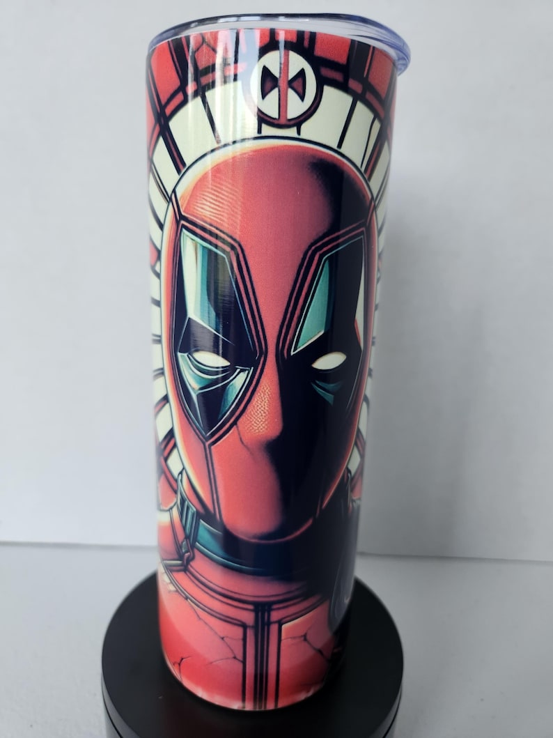 Puede incluir: Un vaso rojo y negro con una ilustraci&oacute;n detallada de la cara de Deadpool. El dise&ntilde;o incluye un patr&oacute;n geom&eacute;trico blanco y rojo alrededor de la parte superior y un logotipo de Deadpool. El vaso tiene una tapa transparente.