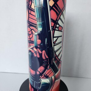 Puede incluir: Un vaso de acero inoxidable con una ilustraci&oacute;n estilo c&oacute;mic. El dise&ntilde;o incluye una figura sosteniendo un arma, sobre un fondo de vidriera. La paleta de colores est&aacute; dominada por el rojo, el negro y el azul.