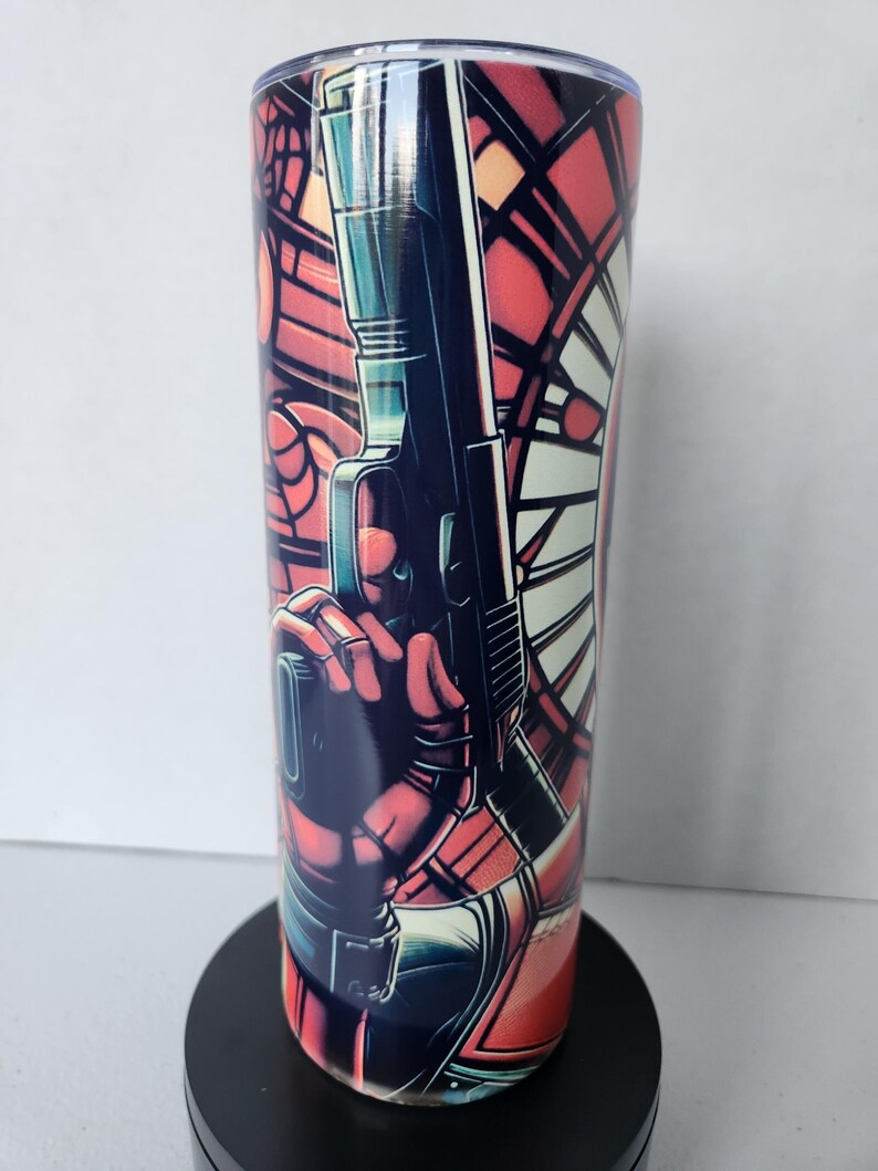 Puede incluir: Un vaso de acero inoxidable con una ilustraci&oacute;n estilo c&oacute;mic. El dise&ntilde;o incluye una figura sosteniendo un arma, sobre un fondo de vidriera. La paleta de colores est&aacute; dominada por el rojo, el negro y el azul.