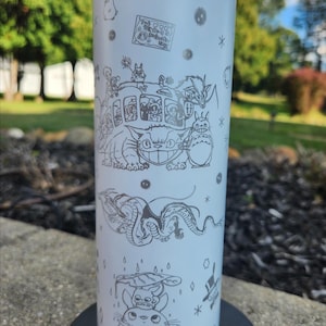 May include: A white, stainless steel tumbler featuring etched illustrations of characters from the animated film "My Neighbor Totoro." The design includes the Catbus, Totoro, and other forest spirits. The tumbler is cylindrical and appears to be approximately 8 inches tall.
