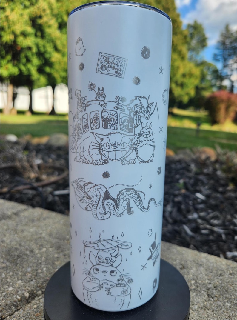 May include: A white, stainless steel tumbler featuring etched illustrations of characters from the animated film "My Neighbor Totoro." The design includes the Catbus, Totoro, and other forest spirits. The tumbler is cylindrical and appears to be approximately 8 inches tall.
