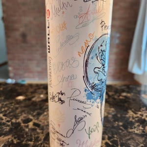 May include: A white insulated tumbler with signatures of characters like Olaf, Cinderella, and Ariel in various colours. The tumbler has a blue and white design and is suitable for hot and cold drinks.