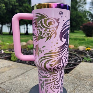 May include: A light pink insulated tumbler with a large side handle and a clear lid featuring a pink flip-top closure. The exterior displays a metallic iridescent rim and a dark purple graphic design of a mermaid and fish with intricate patterns.