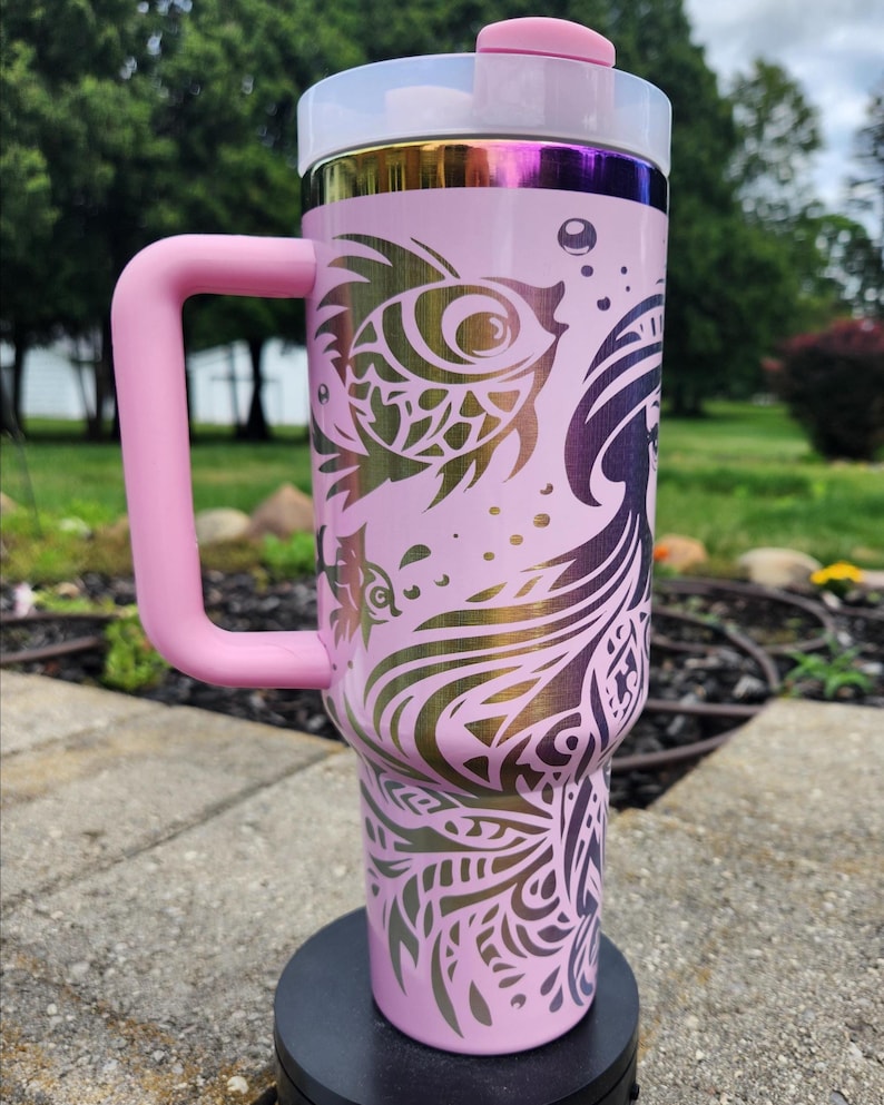 May include: A light pink insulated tumbler with a large side handle and a clear lid featuring a pink flip-top closure. The exterior displays a metallic iridescent rim and a dark purple graphic design of a mermaid and fish with intricate patterns.