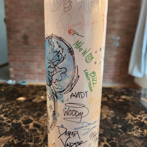 May include: A white stainless steel tumbler with character signatures and illustrations. The design includes names like "Woody," "Darth Vader," and "Buzz Lightyear." The tumbler has a clear lid and is suitable for cold drinks.
