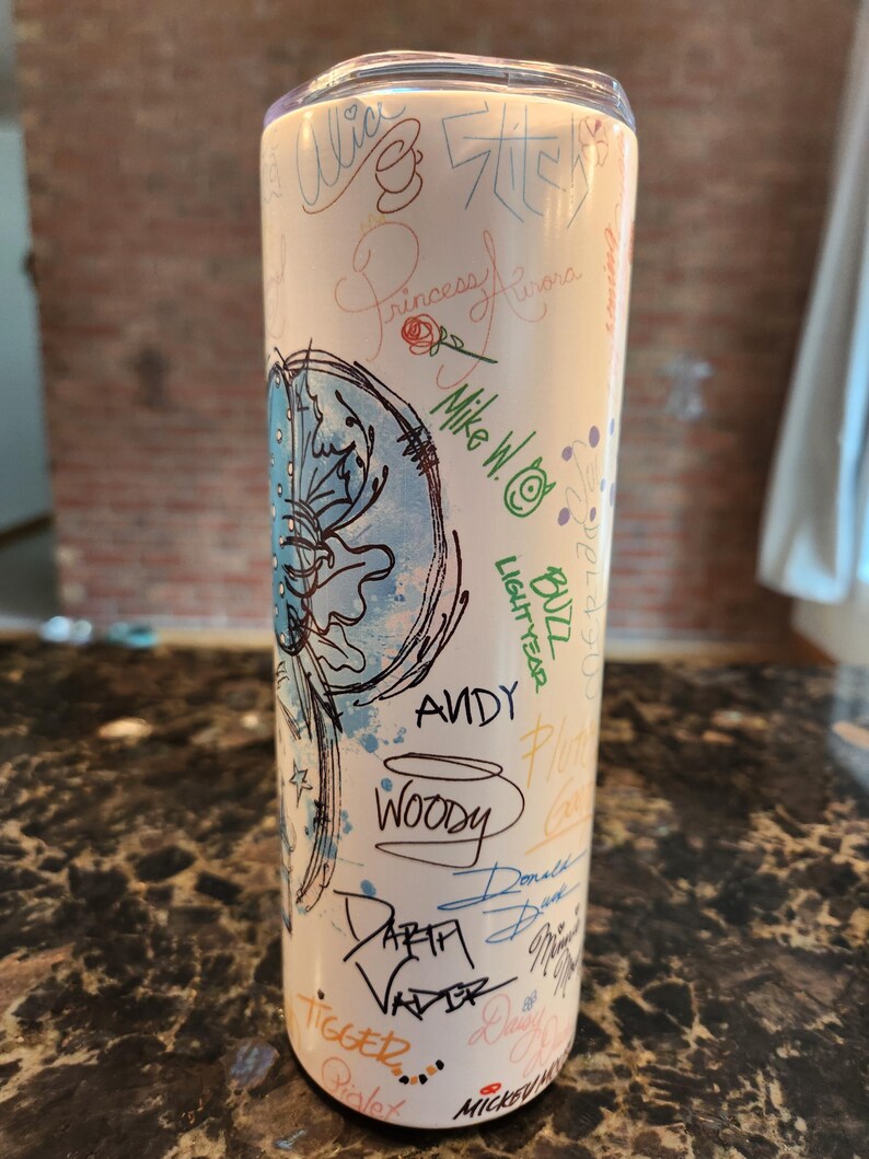 May include: A white stainless steel tumbler with character signatures and illustrations. The design includes names like "Woody," "Darth Vader," and "Buzz Lightyear." The tumbler has a clear lid and is suitable for cold drinks.