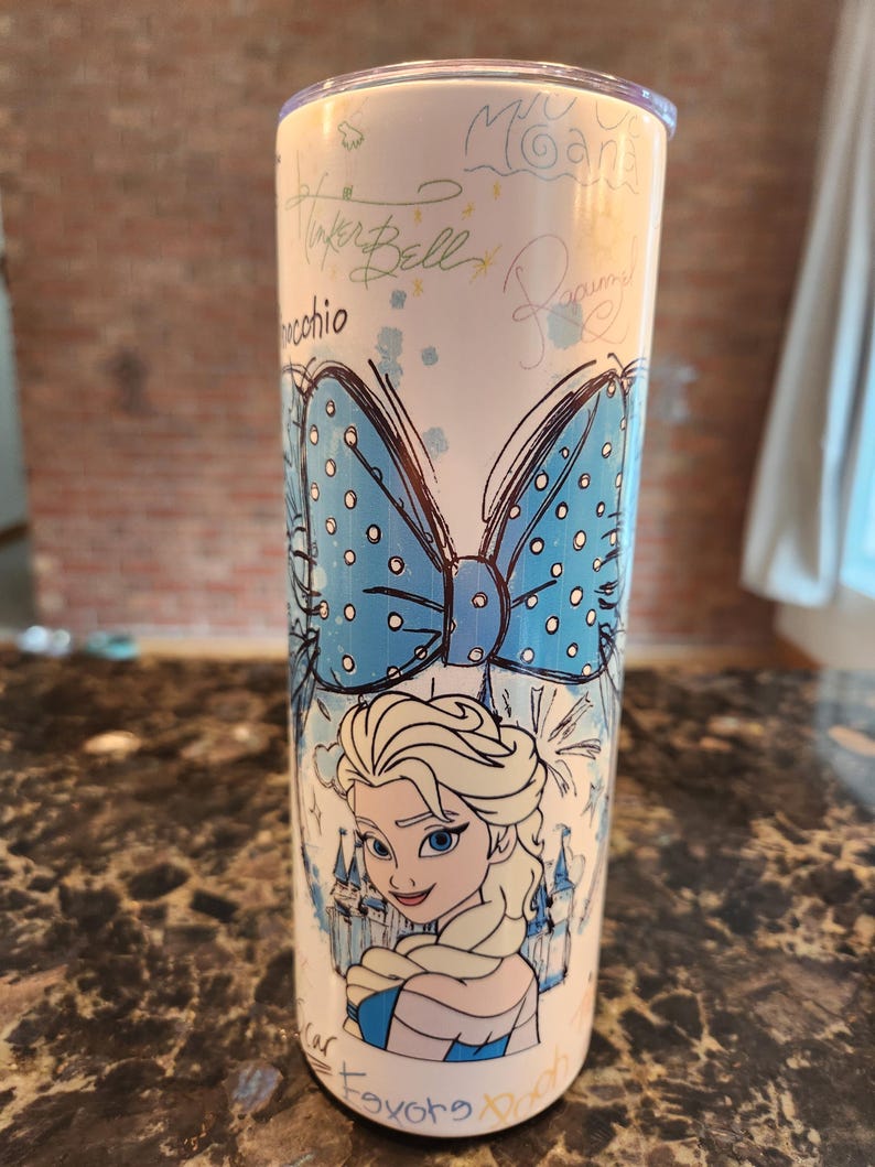 May include: A white tumbler with a cartoon illustration of Elsa from Frozen, a large blue bow, and castle details. The design includes the names of Disney characters like Tinker Bell and Pinocchio. The tumbler has a silver lid.