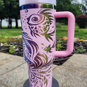 May include: A light pink insulated tumbler with a large handle and a white lid featuring a pink flip-top closure. The exterior is decorated with a dark, iridescent, multi-coloured graphic print of stylised fish and swirling aquatic patterns.