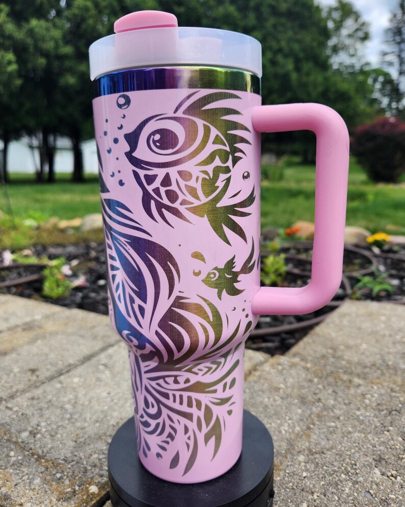 May include: A light pink insulated tumbler with a large handle and a white lid featuring a pink flip-top closure. The exterior is decorated with a dark, iridescent, multi-coloured graphic print of stylised fish and swirling aquatic patterns.
