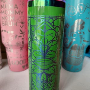 May include: A green insulated tumbler with a decorative blue floral design. The tumbler has a silver rim and a clear lid. Other tumblers in pink and teal are visible in the background.