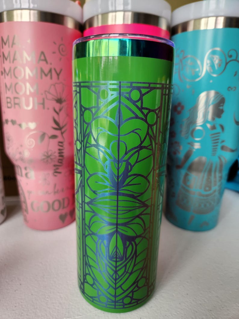 May include: A green insulated tumbler with a decorative blue floral design. The tumbler has a silver rim and a clear lid. Other tumblers in pink and teal are visible in the background.