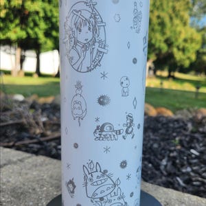 May include: A white tumbler featuring etched illustrations of characters from a popular animated film. The design includes a girl, Totoro, and other whimsical figures. The tumbler is cylindrical and appears to be made of stainless steel.