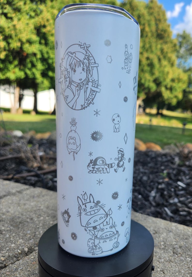 May include: A white tumbler featuring etched illustrations of characters from a popular animated film. The design includes a girl, Totoro, and other whimsical figures. The tumbler is cylindrical and appears to be made of stainless steel.