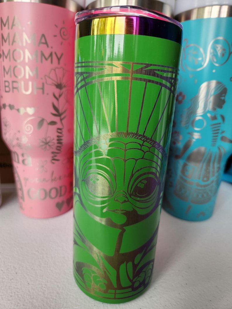 May include: A green stainless steel tumbler featuring a gold and purple graphic of a character's face. The tumbler has a gold rim and a silver lid. Other tumblers in pink and teal are visible in the background.