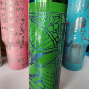 May include: A green tumbler with a geometric design in blue. The tumbler has a silver rim and a pink accent. Other tumblers in pink and blue are visible in the background.