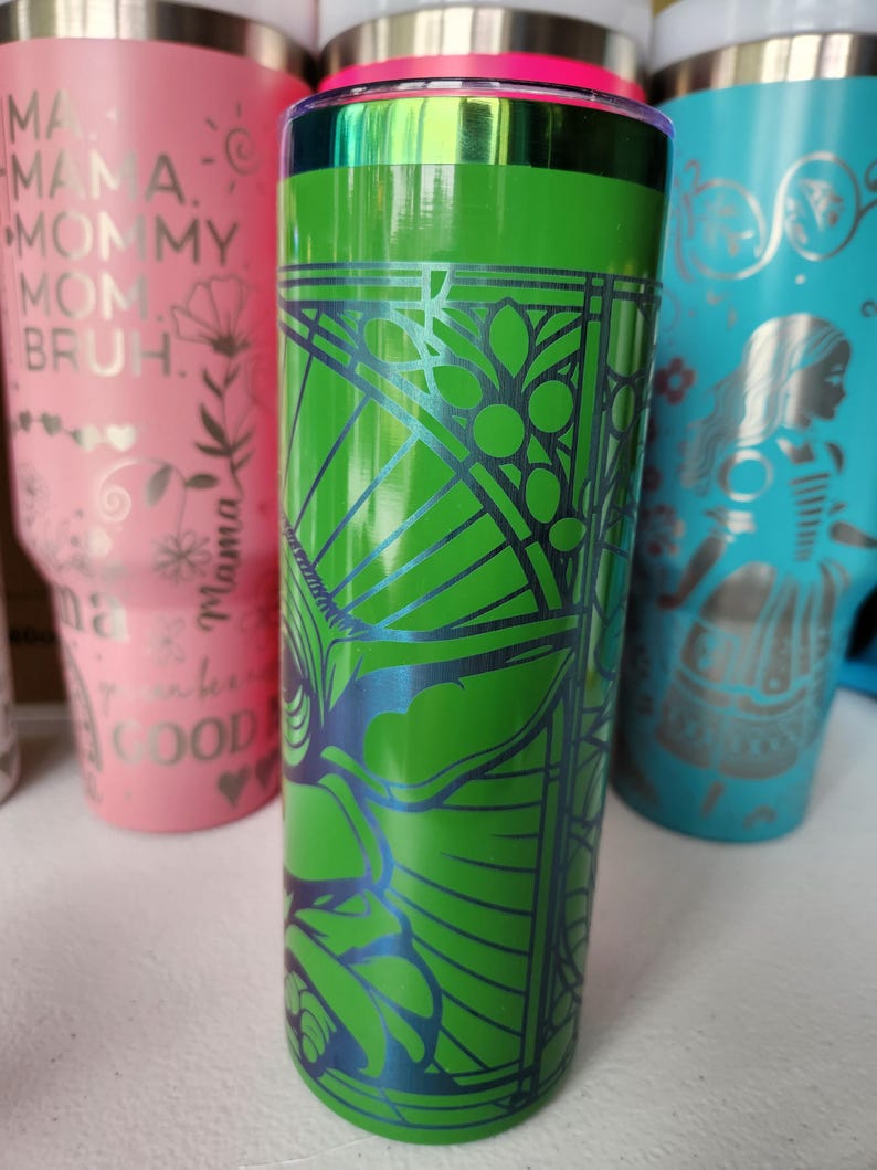 May include: A green tumbler with a geometric design in blue. The tumbler has a silver rim and a pink accent. Other tumblers in pink and blue are visible in the background.