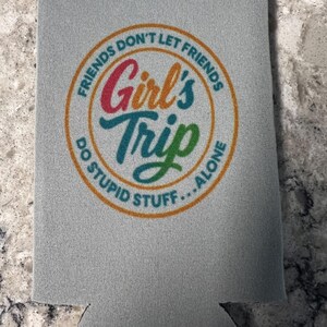 Op de afbeelding: Een grijze blikjeskoeler met de tekst "FRIENDS DON'T LET FRIENDS DO STUPID STUFF...ALONE" in een cirkelvormig ontwerp. De woorden "Girl's Trip" staan in het midden, met een kleurverloop.