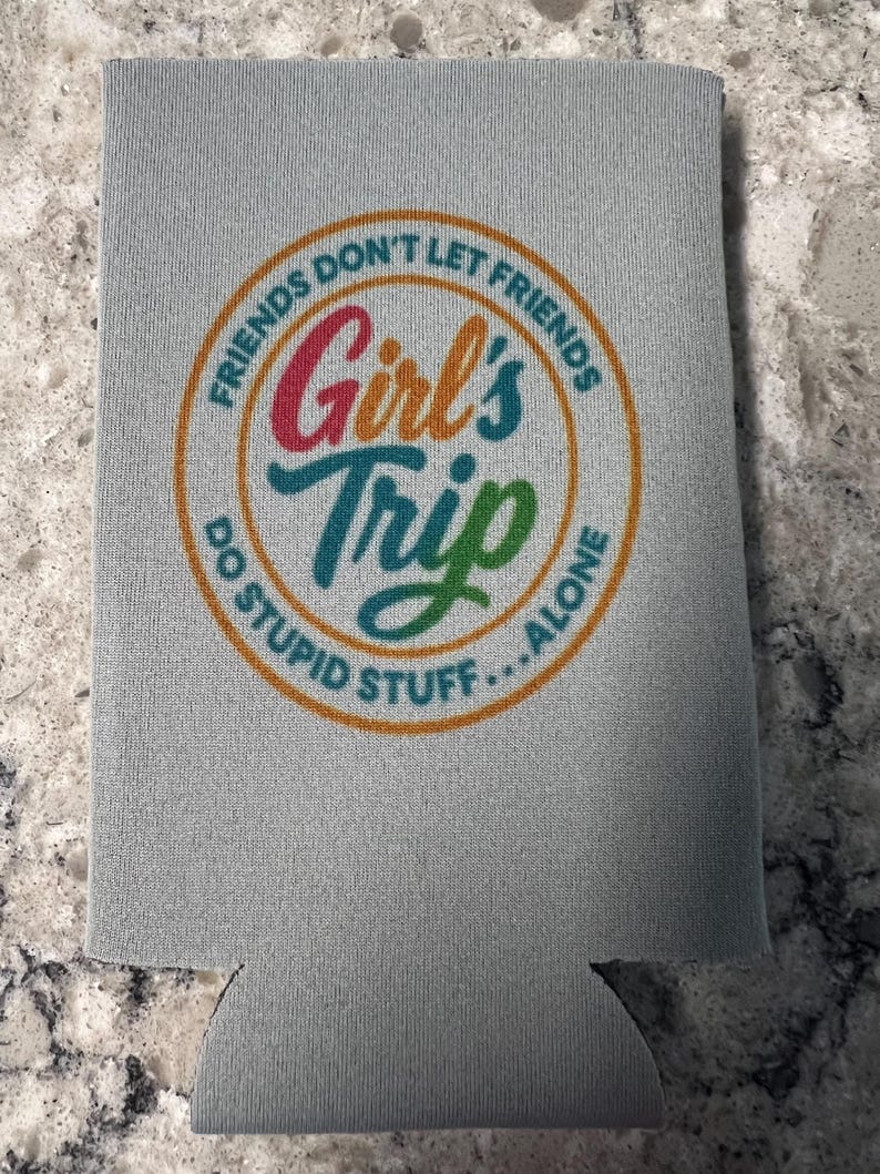 Op de afbeelding: Een grijze blikjeskoeler met de tekst "FRIENDS DON'T LET FRIENDS DO STUPID STUFF...ALONE" in een cirkelvormig ontwerp. De woorden "Girl's Trip" staan in het midden, met een kleurverloop.