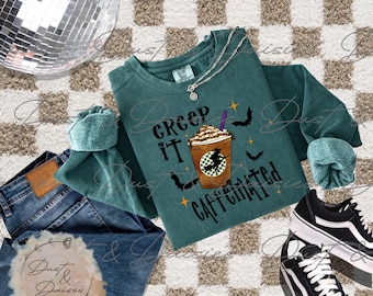 May include: A dark green long-sleeve sweatshirt featuring a graphic of a coffee cup with a witch silhouette, surrounded by bats and stars, with the text "CREEP IT CAFFEINATED." The sweatshirt is paired with blue denim jeans and black sneakers.