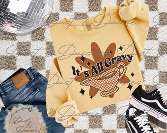 May include: A light yellow crewneck sweatshirt featuring a brown graphic of a gravy boat with a checkered pattern and the text It's All Gravy. The sweatshirt is displayed alongside blue denim jeans and black and white low-top sneakers.