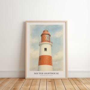 May include: A framed print of Souter Lighthouse, featuring a white and orange striped tower with a red lantern room, set against a cloudy blue sky. The print includes the lighthouse name and location. The artwork is displayed on a wooden floor.