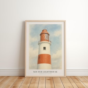 May include: A framed print of Souter Lighthouse, featuring a white and orange striped tower with a red lantern room, set against a cloudy blue sky. The print includes the lighthouse name and location. The artwork is displayed on a wooden floor.