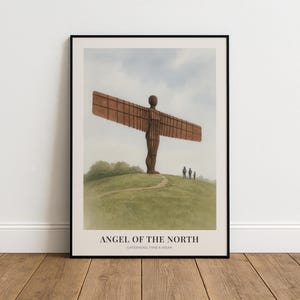 May include: Framed print of the Angel of the North sculpture. The artwork shows the large, rust-coloured angel statue on a grassy hill under a cloudy sky. The print is in a black frame and rests on a wooden floor.