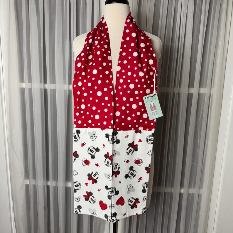 May include: A red and white scarf. The top half has a polka dot pattern, and the bottom half features a Minnie Mouse print with hearts, bows, and flowers. The scarf is displayed on a mannequin.