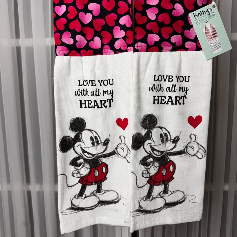 May include: Two white kitchen towels with a black and red heart pattern at the top. The towels feature Mickey Mouse illustrations and the text "LOVE YOU with all my HEART". A tag from "Kathy's Kitchen Boa" is attached.