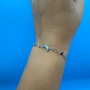 May include: A silver bracelet featuring a crescent moon and phases of the moon design. The bracelet is adorned with small, round, and oval-shaped charms. The bracelet is on a wrist against a blue background.