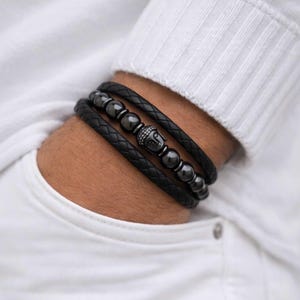 May include: Black leather wrap bracelet with a braided design. The bracelet features dark gray beads and a silver-colored Buddha head charm. The bracelet is wrapped around the wrist.