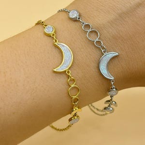 May include: Two delicate bracelets featuring crescent moon charms. One is gold-toned, the other silver-toned. Both have iridescent moon pendants and small, round accents. The bracelets are connected by fine chains.