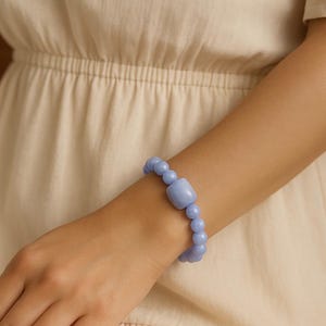 May include: A light blue beaded bracelet featuring a large, square-shaped bead and smaller round beads. The bracelet is worn on a wrist, with a neutral-colored dress in the background. The bracelet is a soft, pastel blue.
