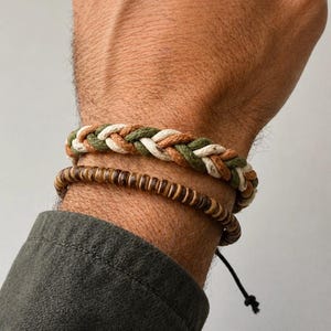 May include: Two bracelets are shown on a wrist. The top bracelet is braided with green, tan, and white rope. The bottom bracelet is made of small, round, brown wooden beads. The wrist is wearing a dark green shirt.