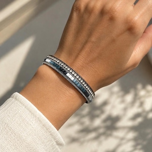 May include: A multi-layered bracelet featuring a central row of dark blue rectangular tiles flanked by two rows of small clear crystals and finished with thin silver-toned metal chains along the edges, creating a structured and layered appearance.